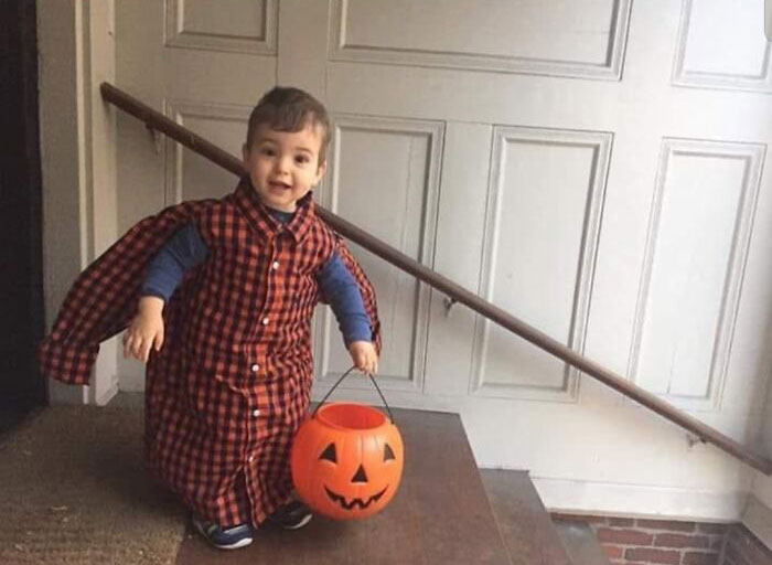 Toddler wearing a long plaid shirt as a cape, holding a pumpkin bucket, capturing kids making parents laugh during Halloween.