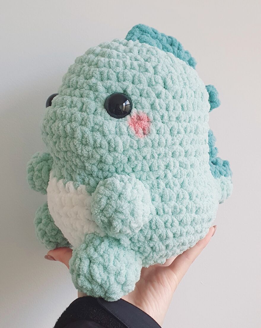 Crochet plush toy in hand, designed to 'cute up' people's days, featuring a soft, fluffy dinosaur with pink cheeks.
