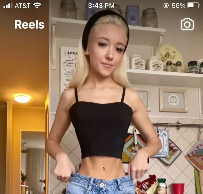 Young woman faking slim waist in Instagram reality post, standing in kitchen while pulling on oversized jeans.