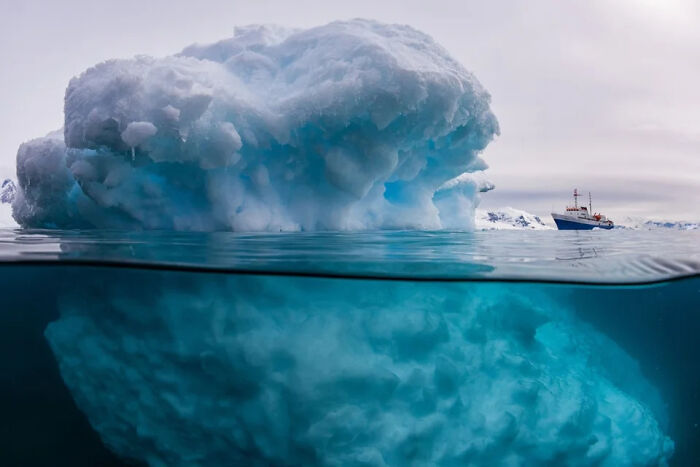 Large iceberg floating in the ocean with a boat nearby, showcasing the vast underwater portion, highlighting ocean dangers.