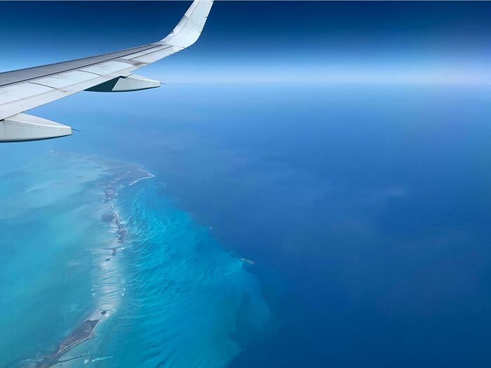 Aerial view of ocean with varying shades of blue and a plane wing, highlighting the vast, deep, and foreboding water below.