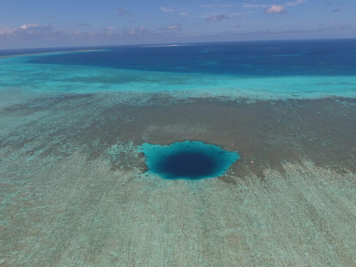Aerial view of a deep blue ocean hole surrounded by shallow turquoise water and coral reefs under a clear sky.