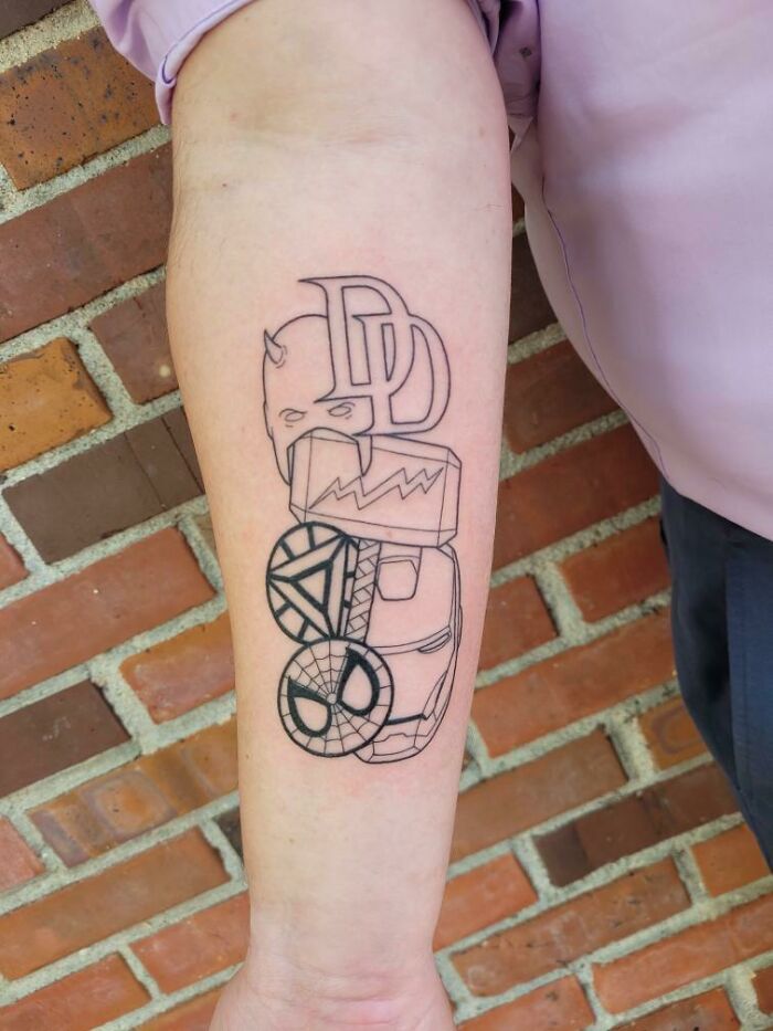 Arm with Marvel tattoos featuring Daredevil, Thor's hammer, Captain America's shield, and Spider-Man mask in black ink.