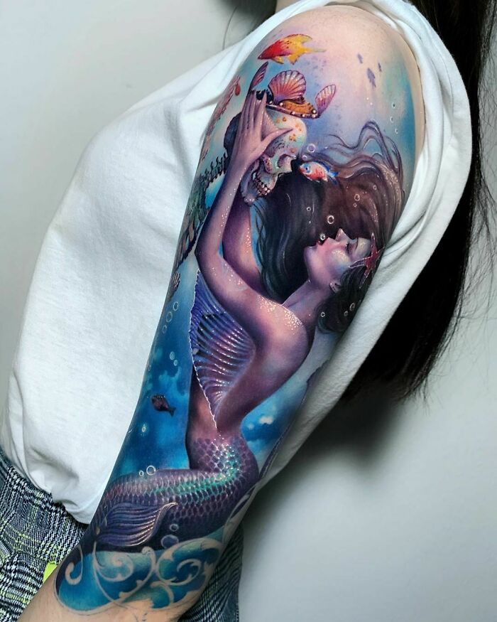 Mermaid tattoo design on arm with skull and fish, showcasing vibrant colors and intricate details.