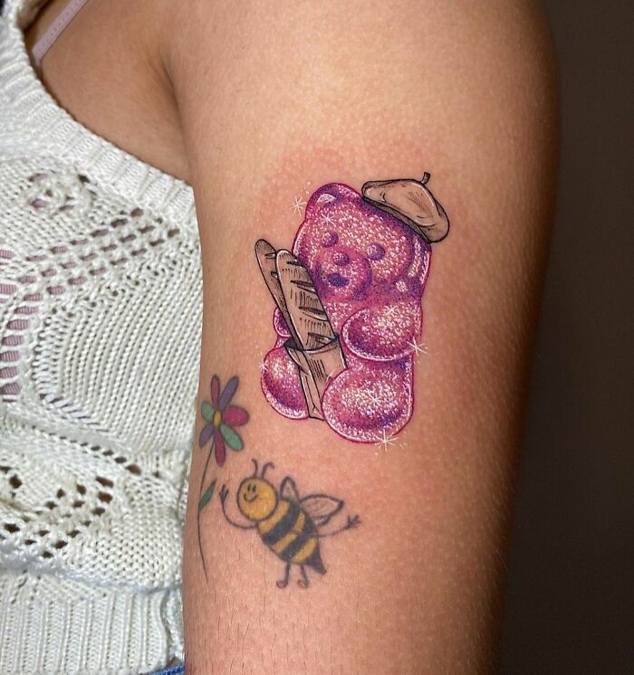 Glitter tattoo of a pink teddy bear with a French theme on an arm, alongside a bee and flower tattoo.