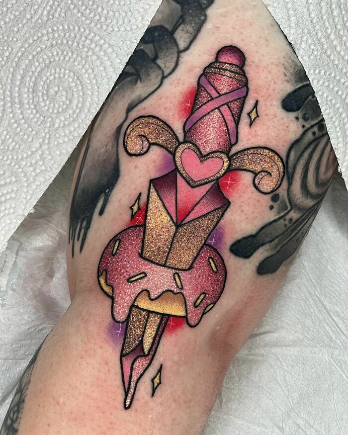 Glitter tattoo of a stylized dagger with a pink donut and heart details on skin.
