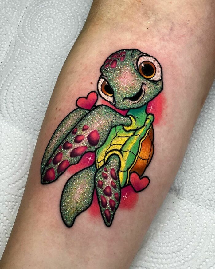A colorful glitter tattoo of a smiling turtle with pink hearts on a person's arm.