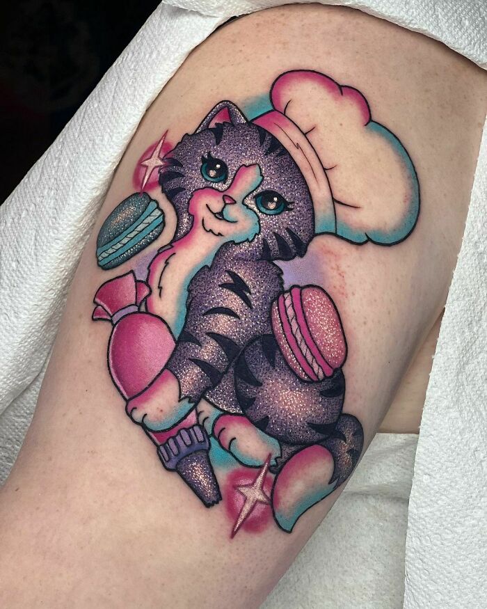 Glitter tattoo of a cartoon cat in a chef hat holding macarons, showcasing vibrant colors and sparkles.