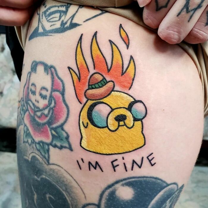 Cartoon tattoo of a yellow dog with flames and "I'm Fine" text, inspired by animated shows, on forearm.