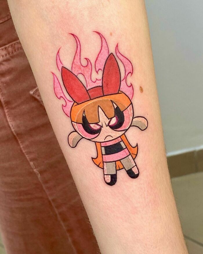 Cartoon tattoo of a character with red eyes and orange hair on a forearm, with pink flames in the background.