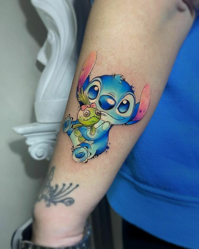 Cartoon tattoo of a blue character holding a green doll on forearm, showcasing childhood favorites.