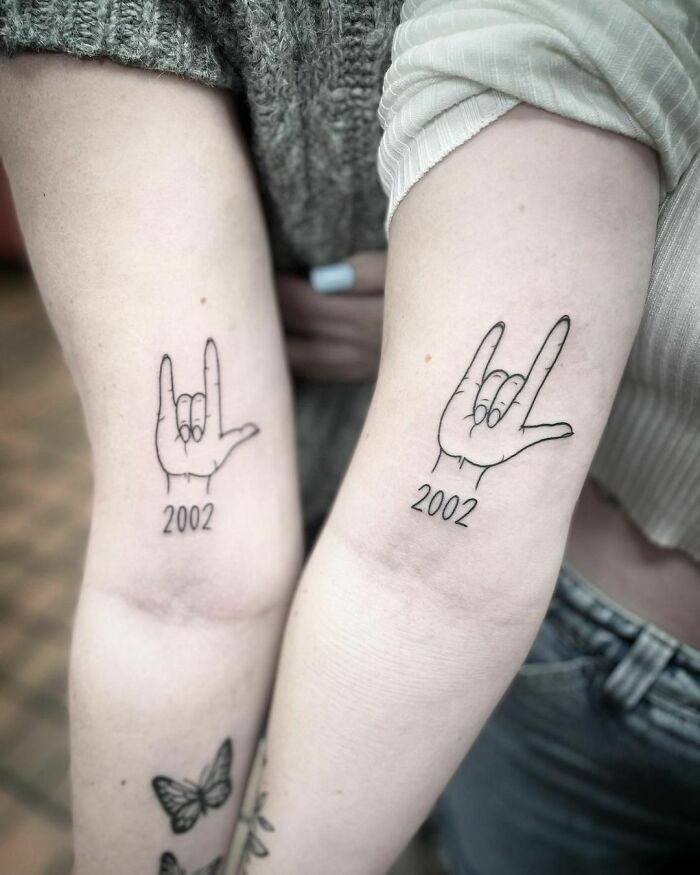 Matching friend tattoos with hand signs and "2002" on forearms, symbolizing friendship.