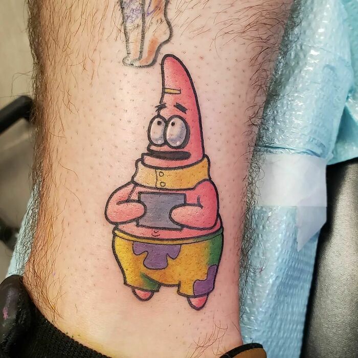 Cartoon tattoo of a pink starfish character Patrick Star on a person's leg, wearing colorful shorts and a yellow collar.
