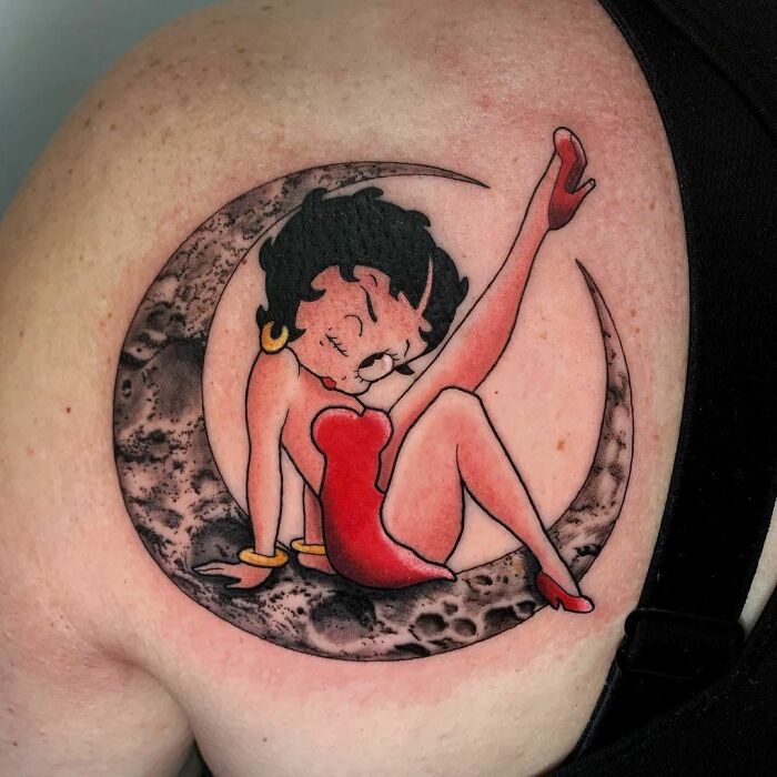 Cartoon tattoo on shoulder featuring a classic character in a red dress sitting on a crescent moon.