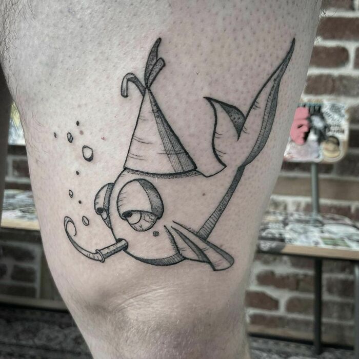 Cartoon tattoos of a whimsical fish wearing a party hat and blowing bubbles, inked on a person's leg.