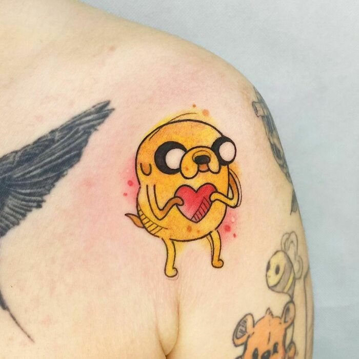 Cartoon tattoo of a yellow dog holding a red heart on a person's shoulder, evoking childhood favorites.