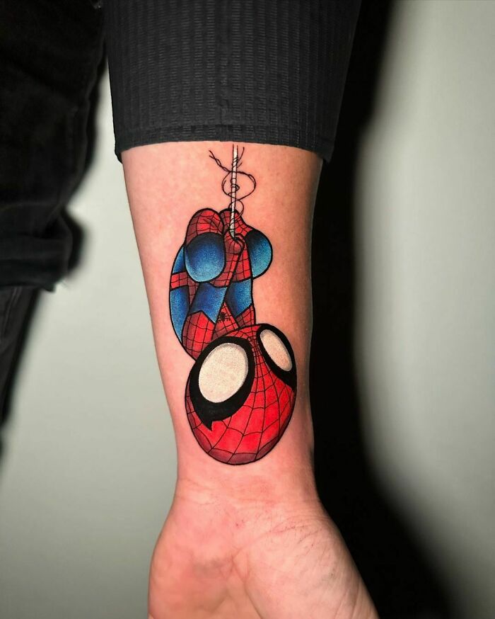 Cartoon tattoo of a superhero Spiderman in red and blue costume on a person's forearm.