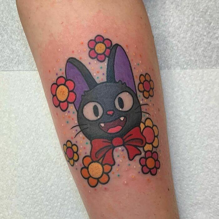 Tattoo of a cartoon black cat with a red bow, surrounded by colorful flowers on an arm.