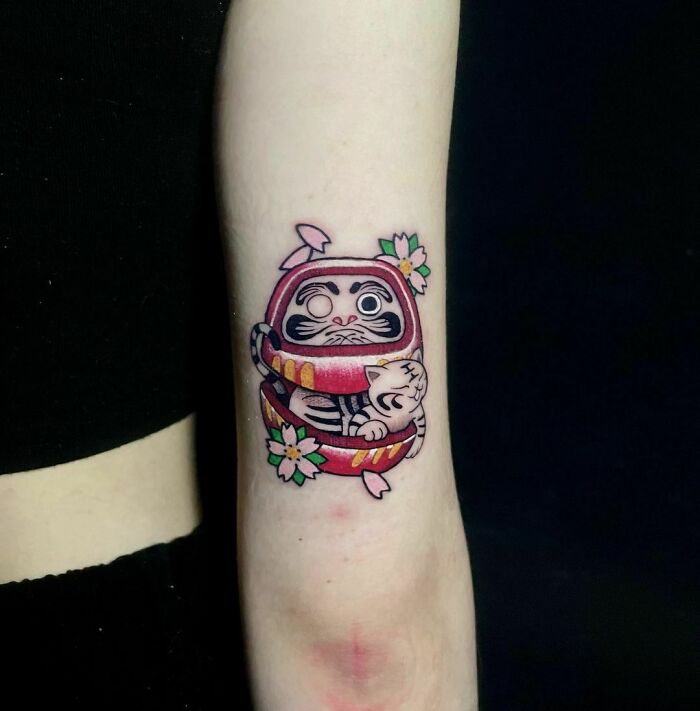 Cartoon tattoo featuring a cute Daruma doll with a cat inside, adorned with pink flowers, on an inner forearm.