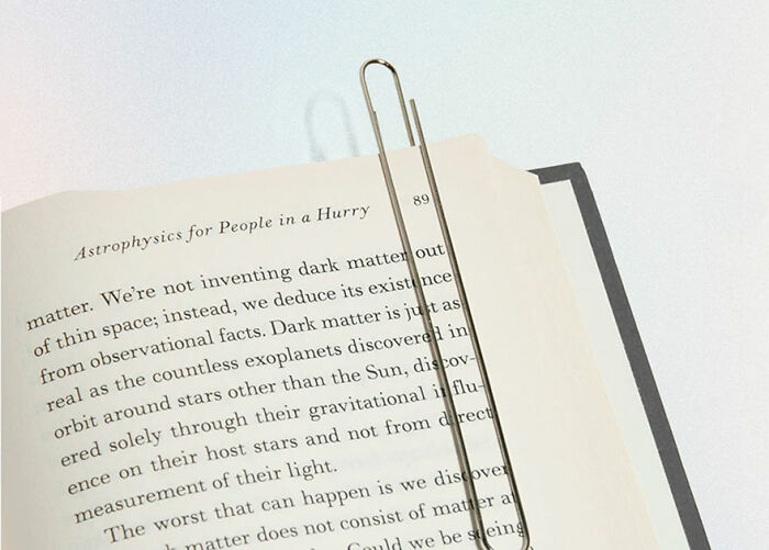 Minimalist product design featuring an elongated paperclip holding an open book page with astrophysics text.