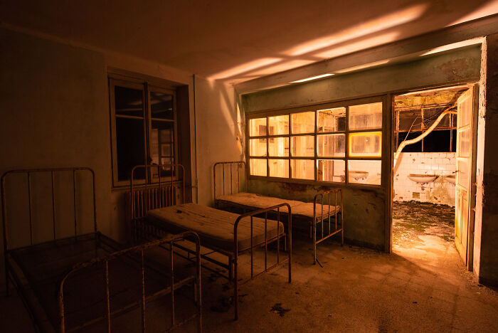 Abandoned place at night with old metal beds, illuminated by warm artificial light through windows and open door.