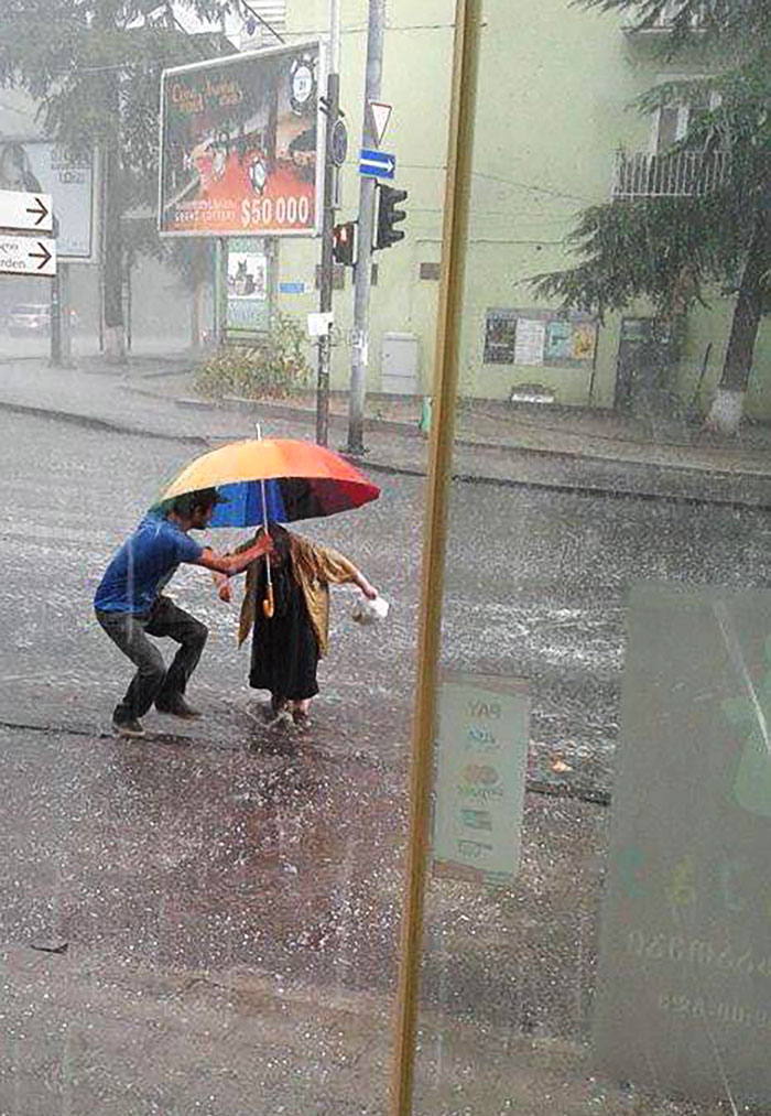A Young Man Sheltering An Old Lady From Hail While Crossing The Road. Tbilisi, Georgia