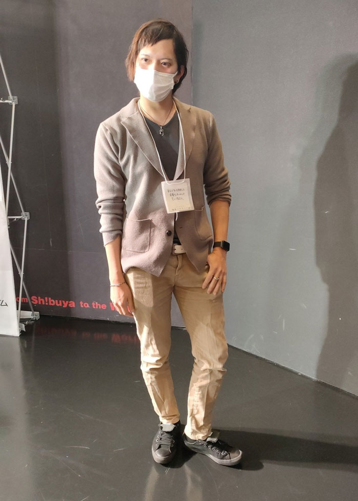 Person in a mundane Halloween costume wearing a mask, cardigan, and ID badge, standing casually against a gray wall.