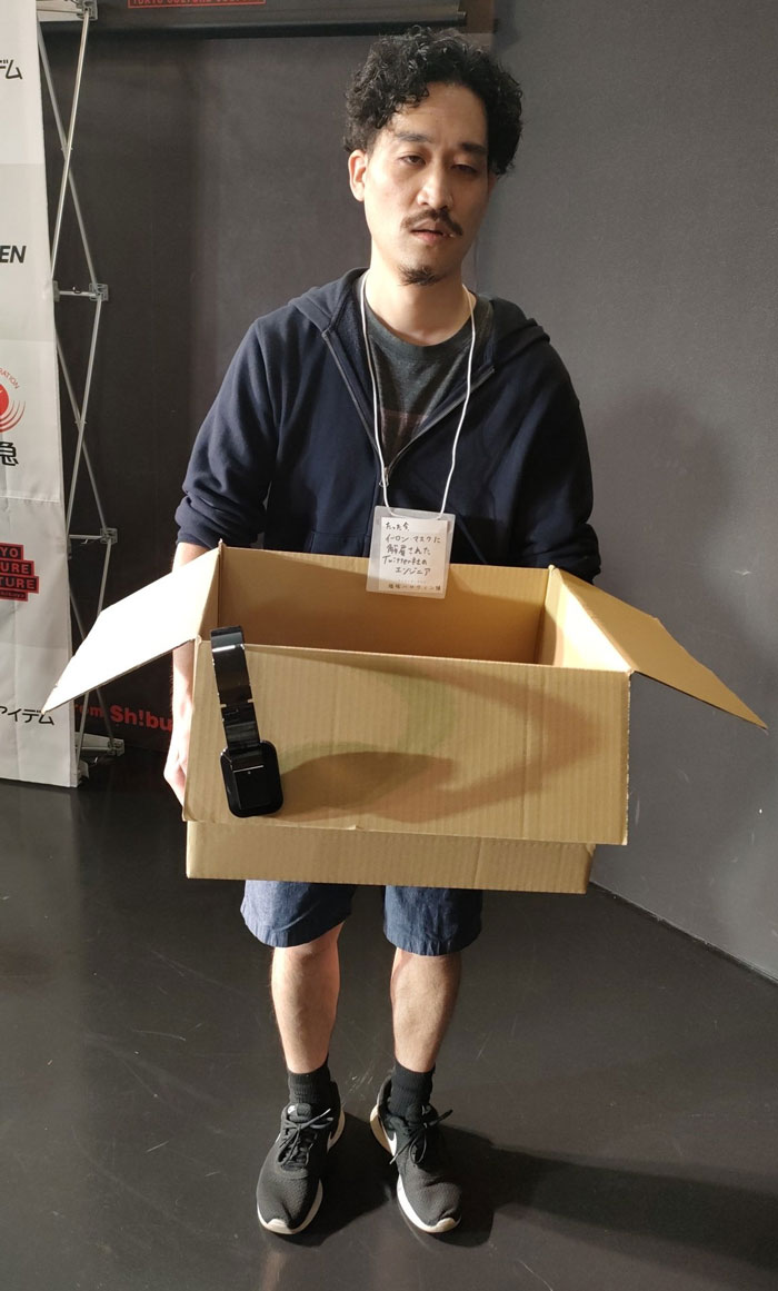 Man in Mundane Halloween costume, holding a cardboard box with a strap, standing indoors.