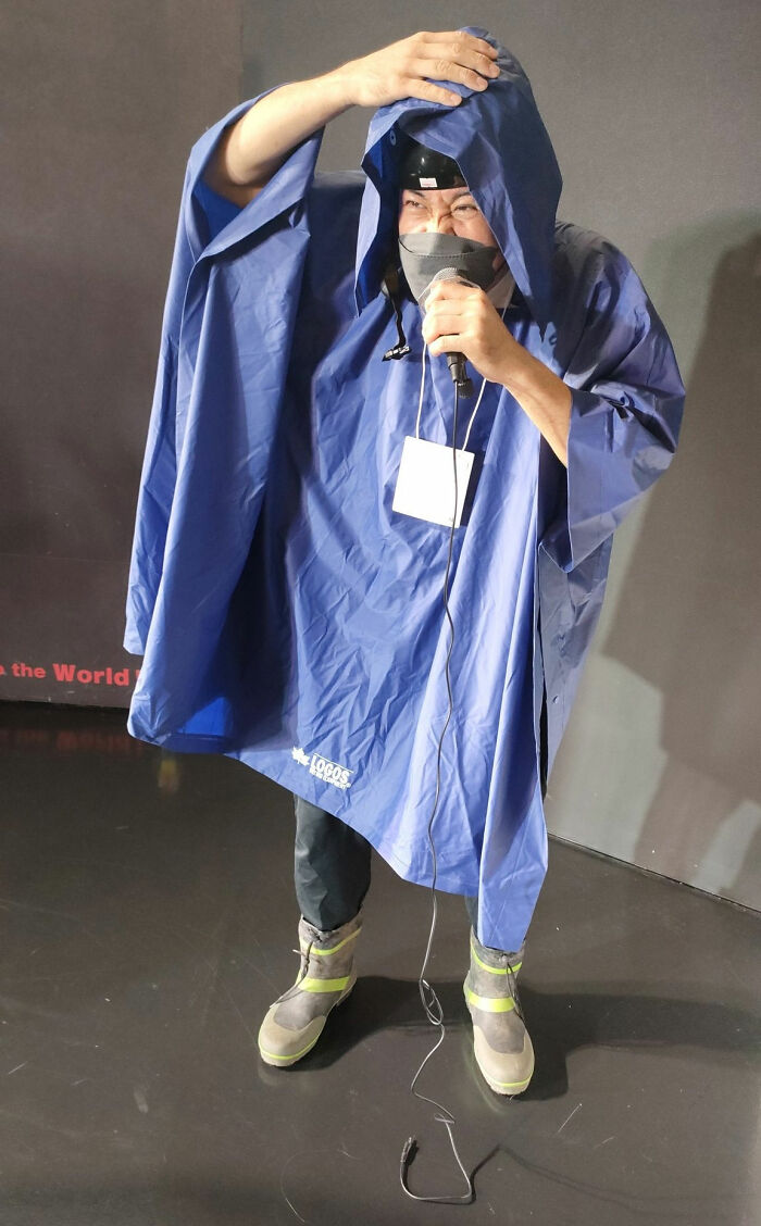 Person in a blue rain poncho with microphone, a quirky "mundane Halloween" costume standing indoors.