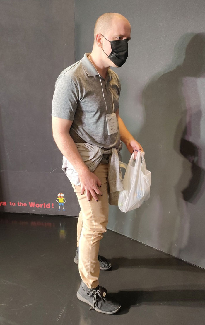 Man in simple costume for Mundane Halloween, wearing a mask and holding a shopping bag, stands against a dark wall.
