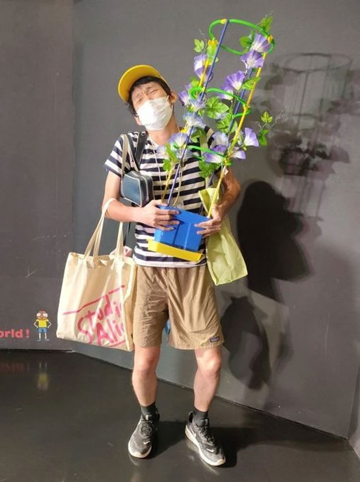Person in mundane Halloween costume holding a potted plant, wearing a striped shirt and yellow cap, standing indoors.