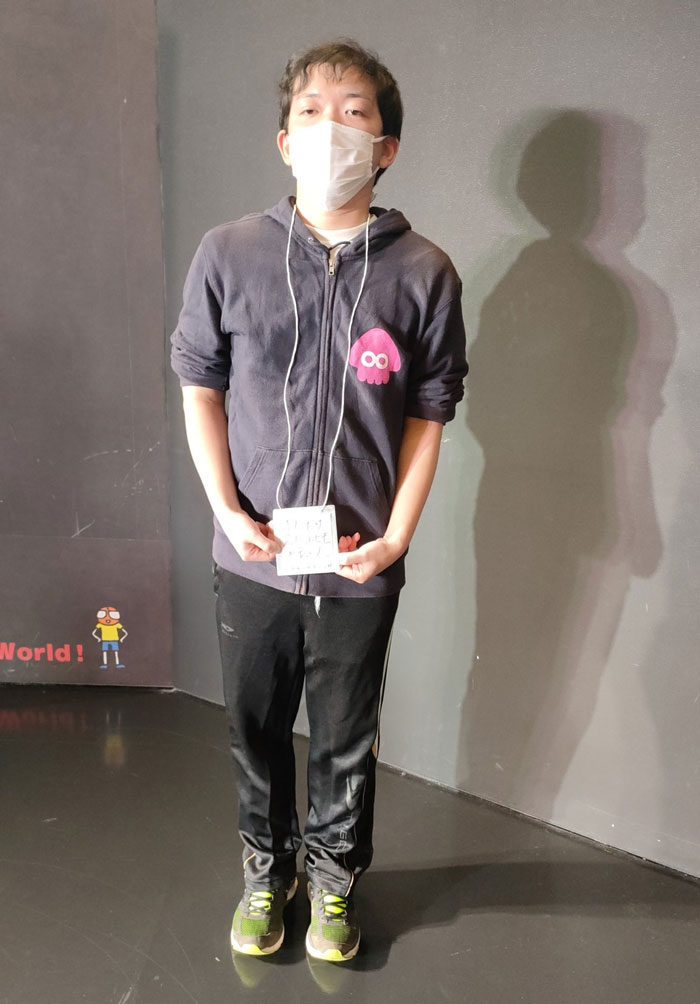 Person in a mundane Halloween costume, wearing a mask and holding a sign, standing against a dark wall.