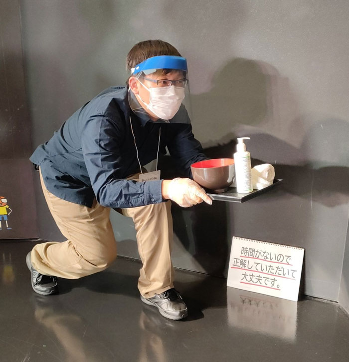 Person in a mundane Halloween costume as a server with face shield, mask, and tray with bowl and sanitizer.