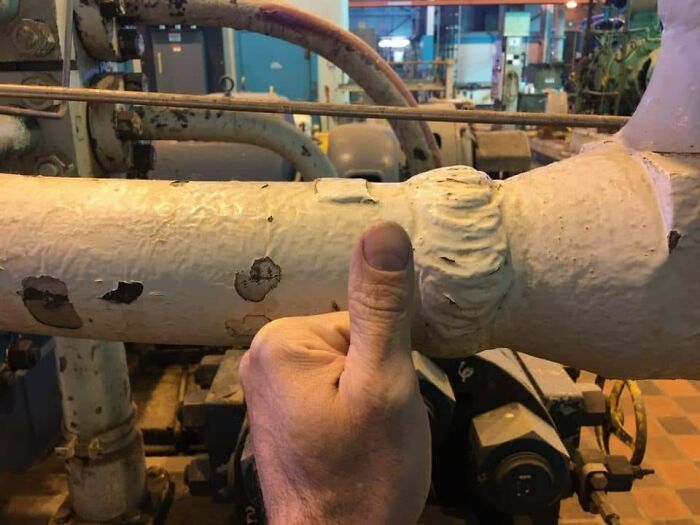 Hand giving a thumbs-up near a chipped white pipe, showing disregard for OSHA safety regulations in an industrial setting.