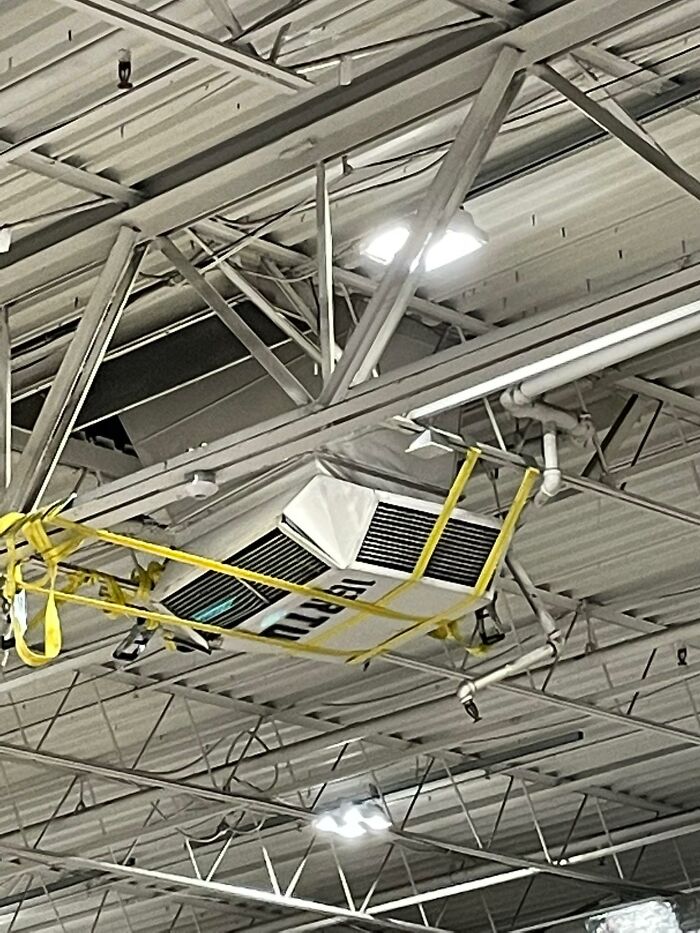 Air conditioning unit in a warehouse ceiling, secured with yellow straps, illustrating lack of OSHA safety regulations adherence.
