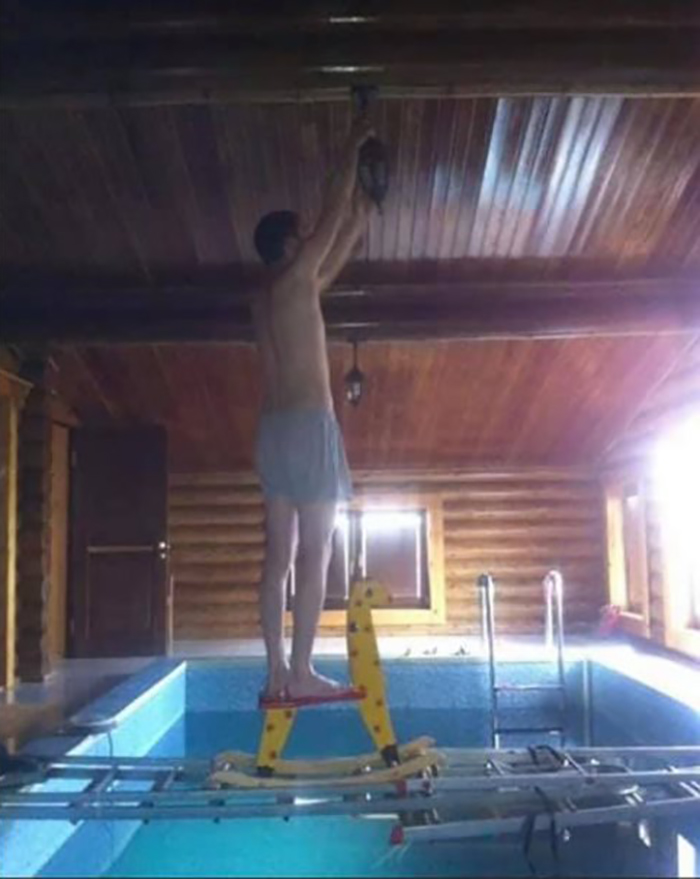 Man balancing on makeshift platform over indoor pool, ignoring OSHA safety regulations.