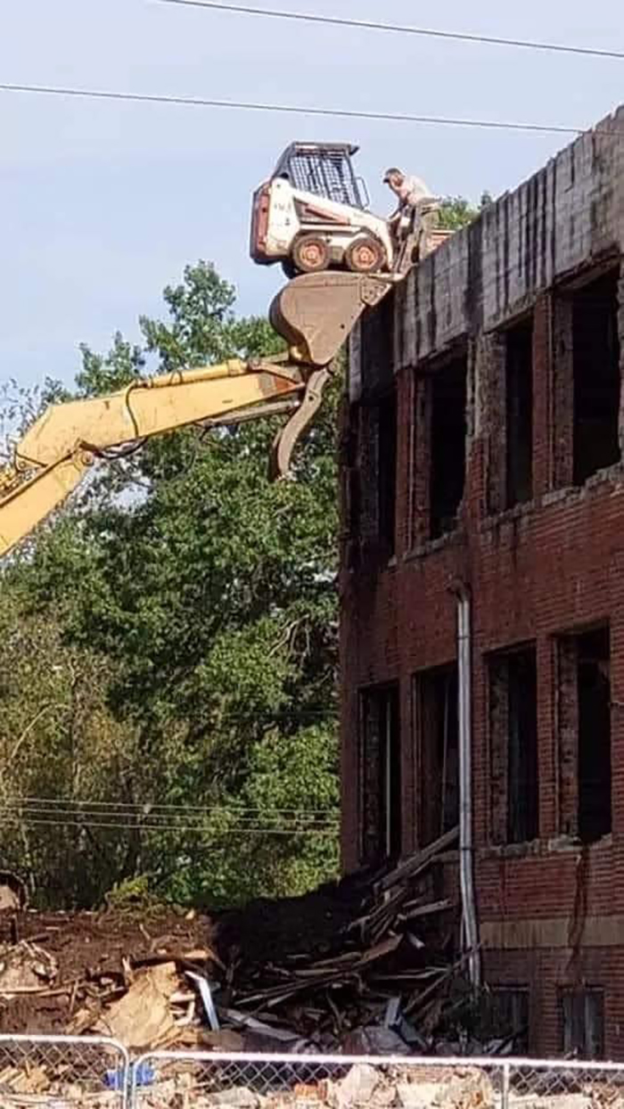 Excavator lifts a skid loader to building roof, ignoring OSHA safety regulations, amid construction site trees and bricks.