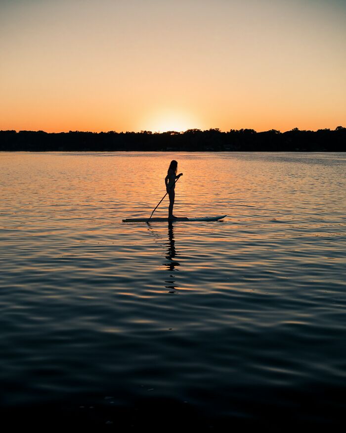 Woman Paddle Boarding In Lake While The Sun Is Setting 
