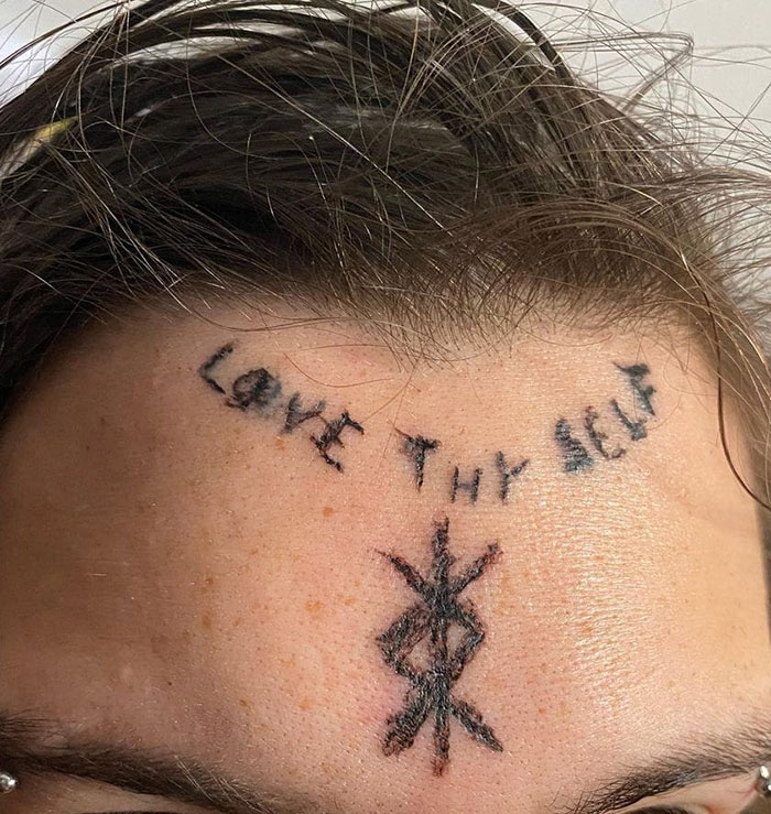 Poorly executed forehead tattoo reads "Love Thy Self" with a black symbol below it.