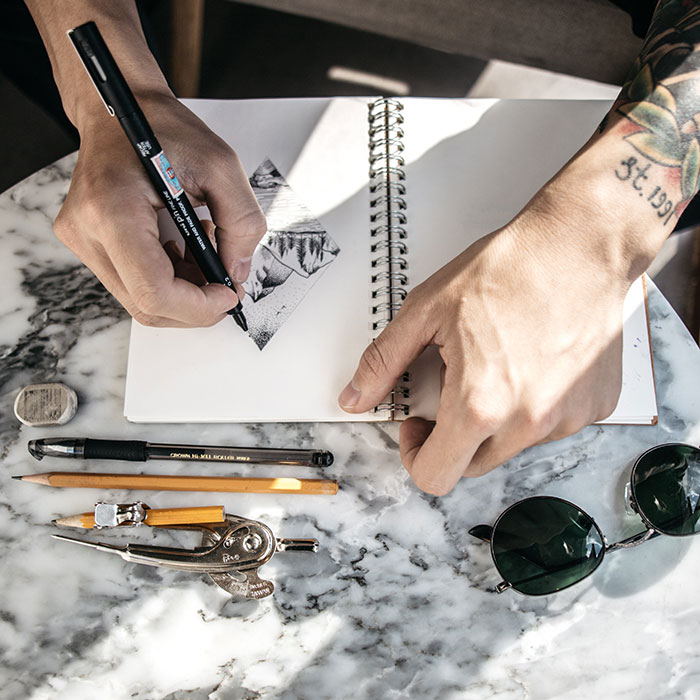 Person sketching alone on a marble table, with drawing tools and sunglasses, emphasizing the keywords "things to do by yourself."