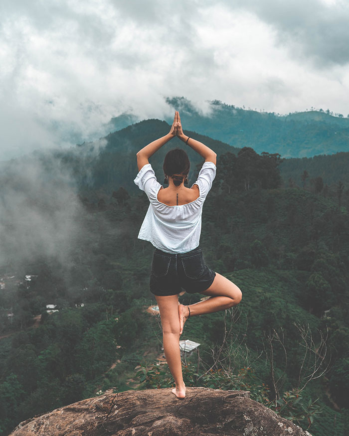 Person practicing yoga alone in nature, standing on a rock with a forest and mountains in the background.