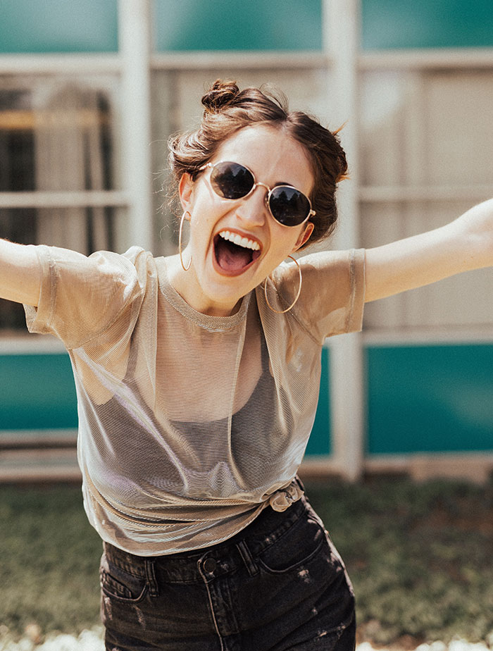 Woman enjoying me time, laughing outdoors, wearing sunglasses and a sheer top.