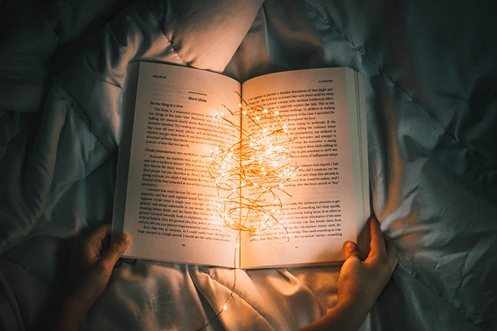 Person enjoying me time, holding a book with glowing fairy lights on a cozy bedspread.