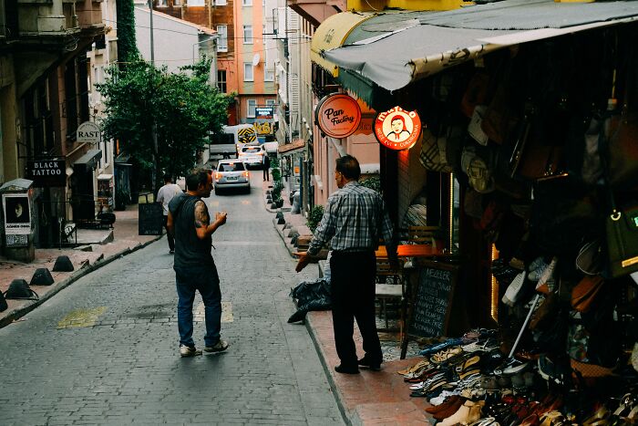 Two men talking on a narrow street lined with shops, highlighting people noticing lack of basic facts awareness.