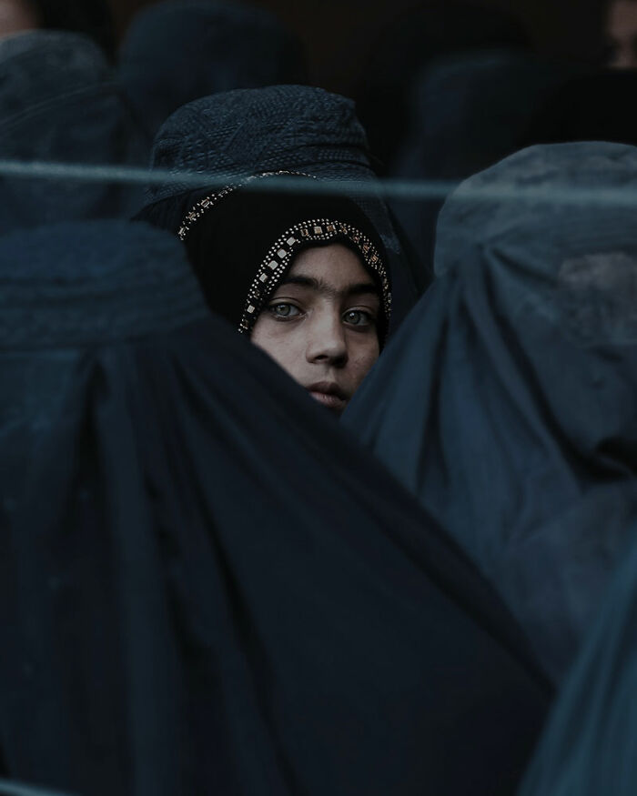 Young Afghan woman with piercing eyes wearing a decorated headscarf among women dressed in dark traditional clothing.