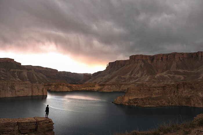 Person standing on rocky cliff overlooking dramatic lake and mountains during a moody sunset while traveling in Afghanistan scenery