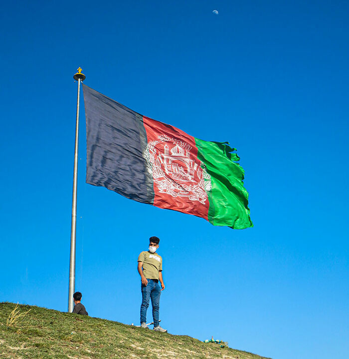 Young man standing near a large Afghanistan flag on a hill, showcasing travel photography in Afghanistan.