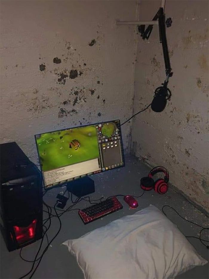 Cursed gaming setup with a floor pillow, red LED PC, monitor, keyboard, mouse, headphones, and wall-mounted microphone.