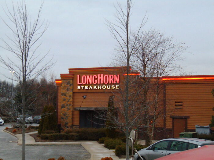 Longhorn Steakhouse restaurant exterior with illuminated sign, leafless trees, and parked cars in a quiet parking lot.