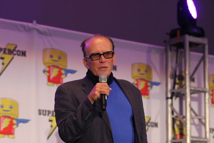 Male celebrity wearing glasses and a blue shirt speaking on stage, showcasing talent and intelligence at an event.